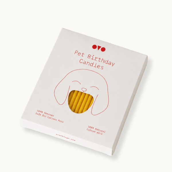 A boxed candle product labeled 'Pet Birthday Candles' shown at an angle on a clean white background. The package is white with a simple red line drawing of a dog face and red text, with a clear window revealing beeswax candles inside.