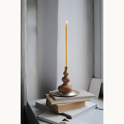 Wooden candle holder with a lit slim beeswax candle on top of books against a light gray wall.