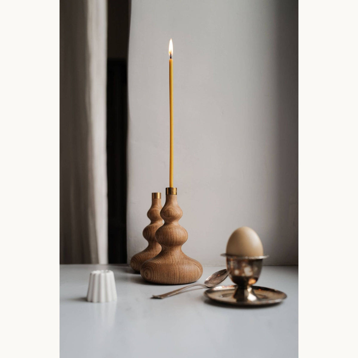 Wooden candle holder with a lit slim beeswax candle, egg cup with an egg, and small white object on a light surface.