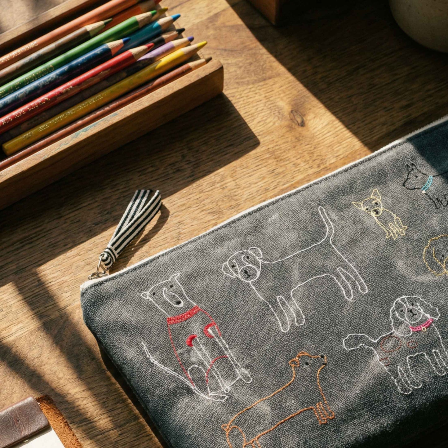 A gray fabric zippered pouch with embroidered dog illustrations, styled on a wooden desk next to a pencil box.