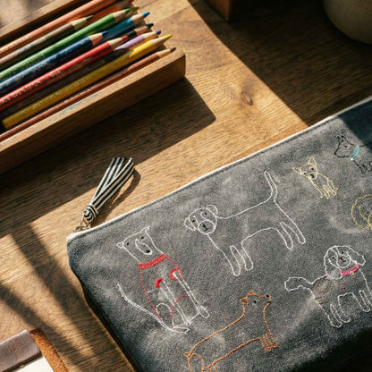 A gray fabric zippered pouch with embroidered dog illustrations, styled on a wooden desk next to a pencil box.