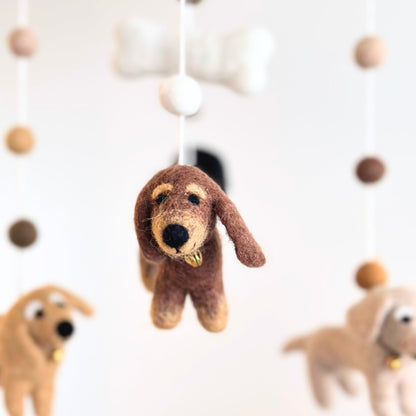 A brown felted dog hanging from a mobile, with other soft plush dog toys blurred in the background against a white wall.