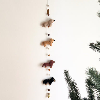 A vertical hanging felt dachshund dog garland with four small felt dog figures in beig, tan, brown, and black, suspended between white and brown beads, shown against a plain white wall with pine branches at the right edge.
